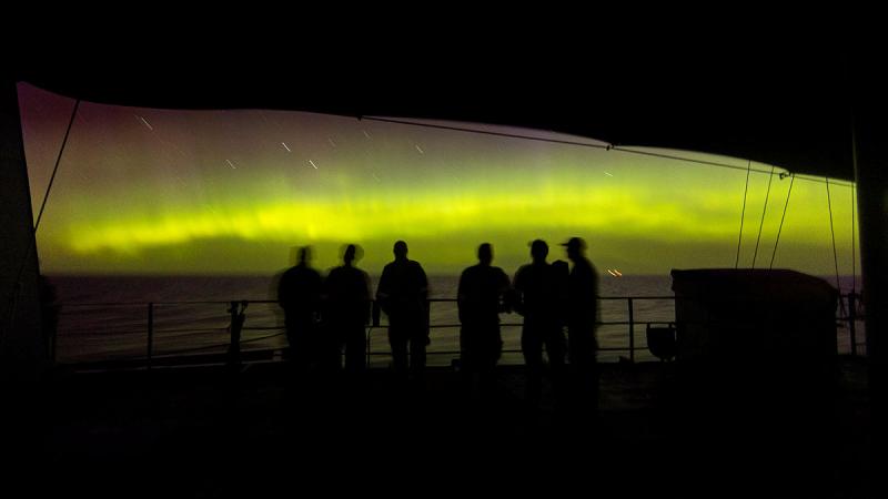Six sailors stand on the deck of a ship and view a yellow night sky caused by the Aurora Australis.