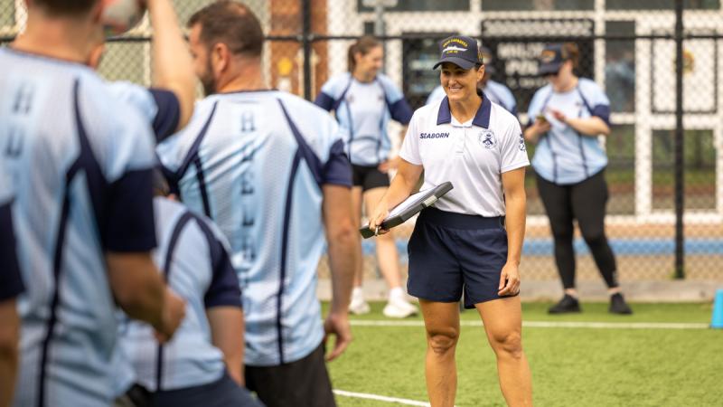 A Navy physical training instructor holds a clipboard and supervises participants on an enclosed AstroTurf field.
