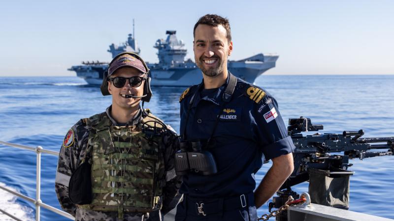 Two men standing on the deck of a ship, with an aircraft carrier in the water behind them.