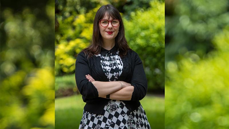A woman wearing red glasses, a black and white dress, and a black cardigan stands in front of greenery.