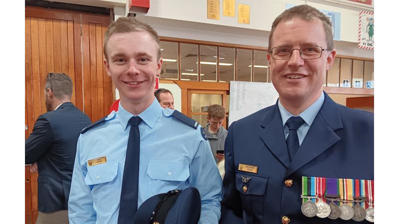 Two men in uniform stand indoors smiling, the older man wearing several medals.