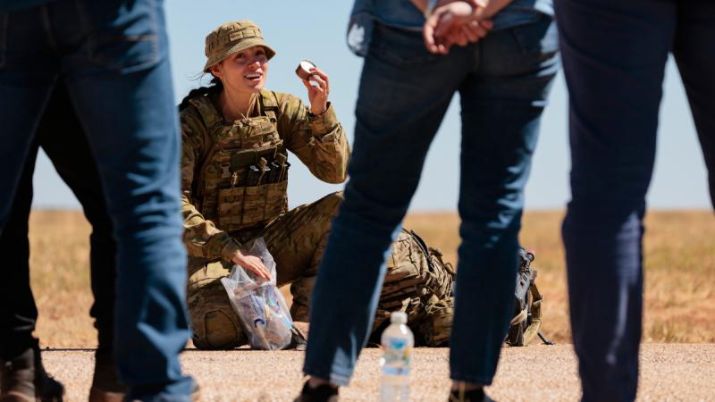 A soldier crouches on the ground displaying ration pack items to several people standing nearby.
