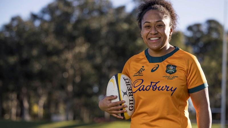 A female rugby player in a gold and green team shirt and holding a rugby ball stands outdoors on a fine day.