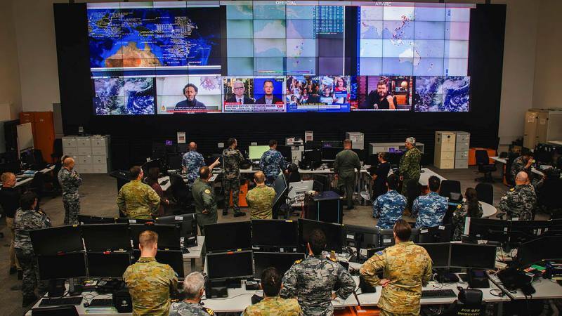 A meeting room is filled with Defence personnel in uniform facing large screens.