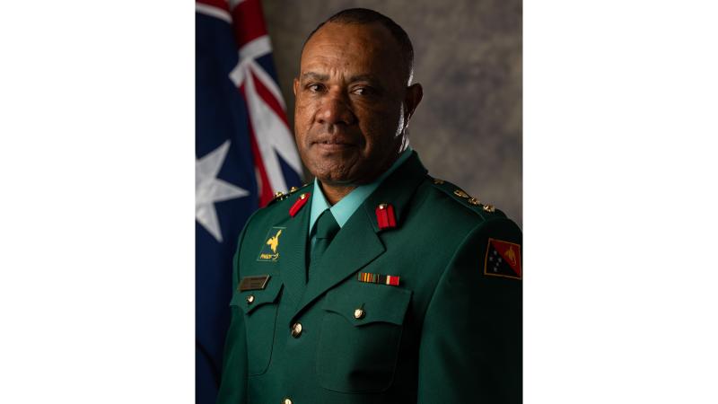 A Papua New Guinean officer in a dark green uniform stands for a photo in front of an Australian flag. 