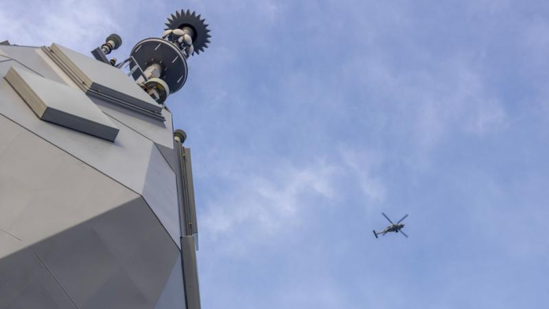 A helicopter flies in the sky with a small section of a ship visible at the edge of the frame.