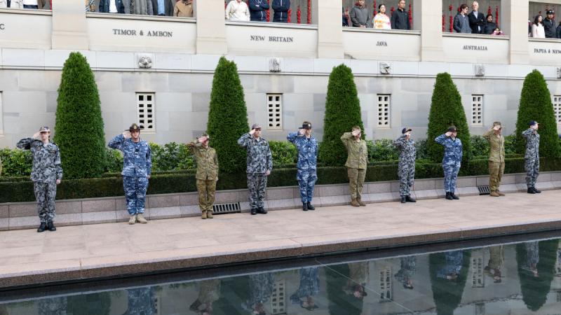 People in military uniform stand and salute beside the memorial pool as people watch from a balcony above.