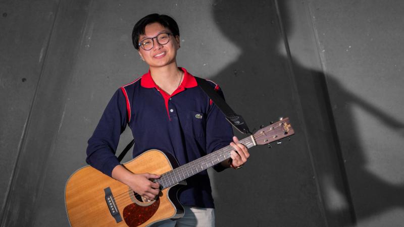 A woman in casual clothes stands in front of a dark grey background holding an acoustic guitar.