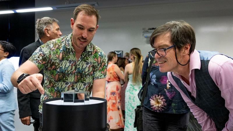 A man in a bright shirt points at a project he is showcasing and explains it to another man who is looking on.