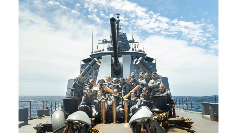 Sailors standing beneath a 5-inch gun on the deck of the ship, some with ammunition held in their hands.
