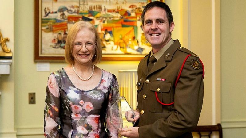 A woman stands beside a man in uniform holding a glass award.