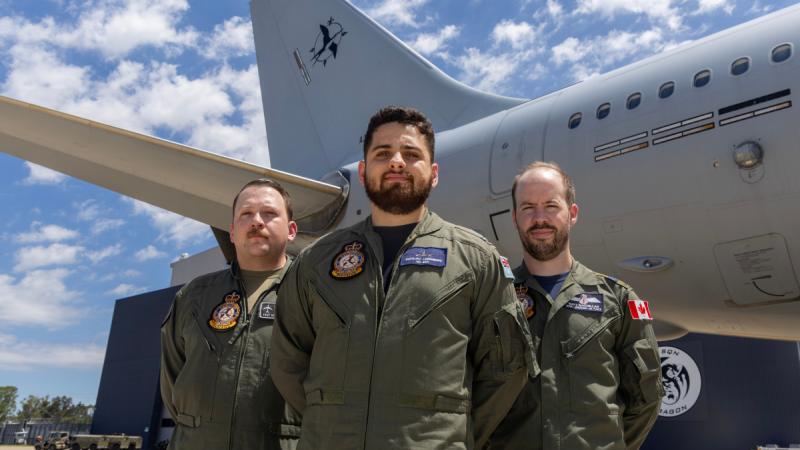 Three men in flight gear stand in front of a large aircraft tail.