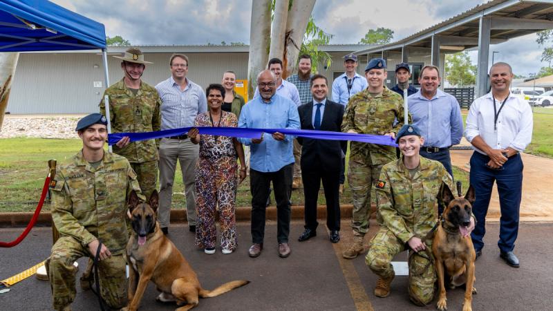 A group of people and two dogs stand in a car park in front of a building while the man in the centre cuts a blue ribbon.