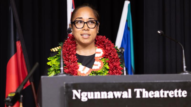 A woman wearing leis talking at a lectern, with the Australian, Aboriginal and Torres Strait Islander flags behind her.