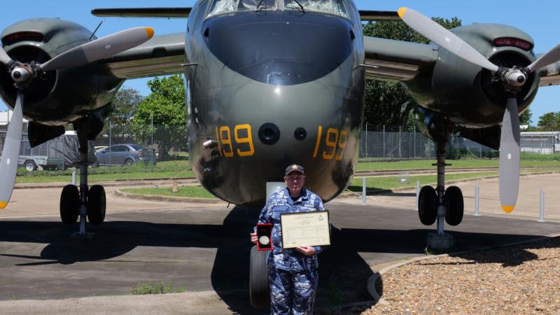A man in Air Force fatigues holds a framed certificate and a medal in a presentation box while he stands in front of a green aircraft with propellers on both wings.