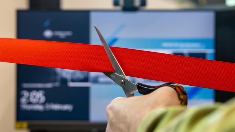 A hand holds scissors cutting red tape in front of a computer screen.