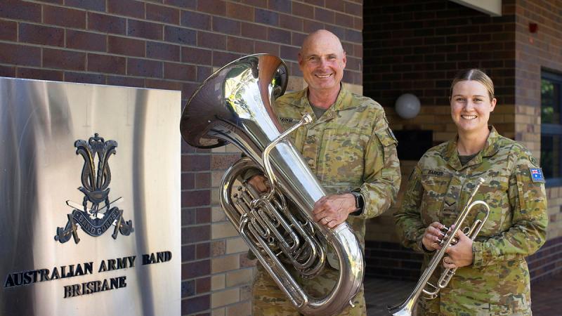 Two Army personnel stand with a trumpet and a tuba in front of an Australian Army Band - Brisbane sign.