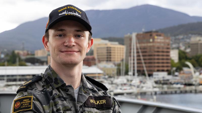 A sailor in Navy fatigues stands on a ship’s deck with a harbour, city skyline and mountains in the background.