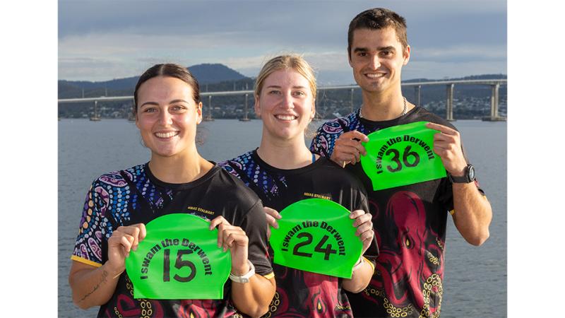 Two women and a man stand before a body of water with green swimming caps.