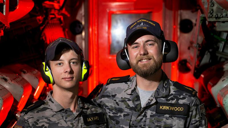 Two Navy personnel in uniform stand in a ship's engine room filled with red light and metal equipment. 