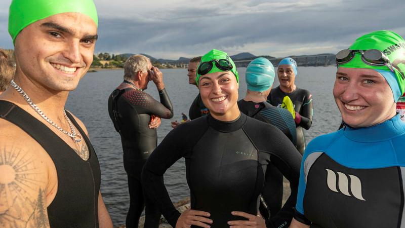 A small group of Navy personnel wearing caps and wetsuits stand near the Derwent River as they prepare to swim in it.