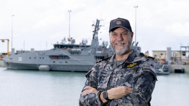 An man in Navy uniform stands onshore in front of a ship.