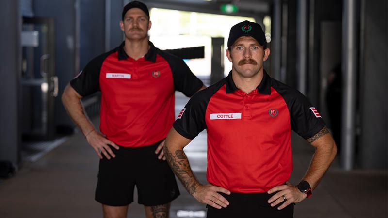 Two men wearing baseball caps and red shirts stand with their hands on their hips.