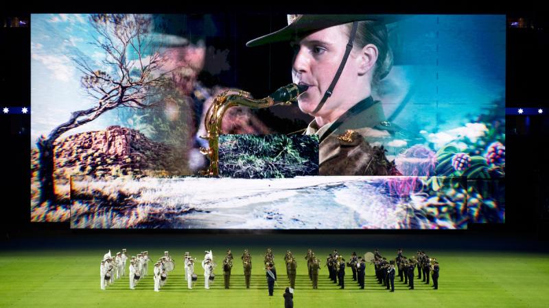 A large screen above a stadium field displays a woman in Army uniform playing a tenor saxophone, with a marching band on the field below.