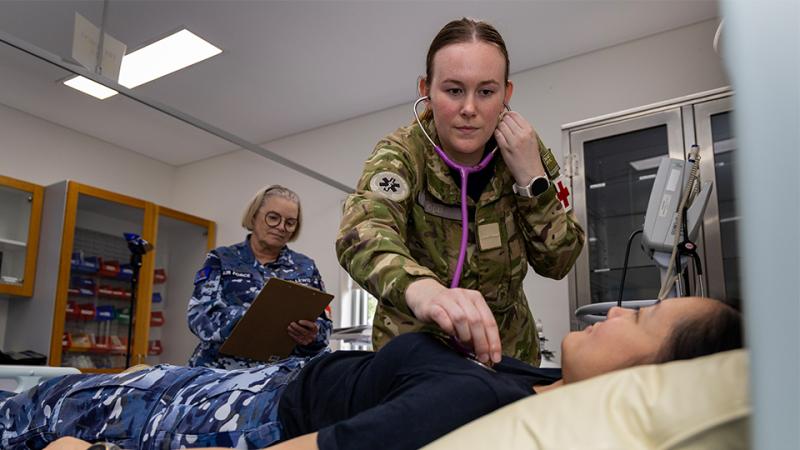 A woman in blue military uniform takes notes in a medical room while another woman in a green military uniform leans towards a patient on a hospital bed to listen to the patient’s chest with a stethoscope.