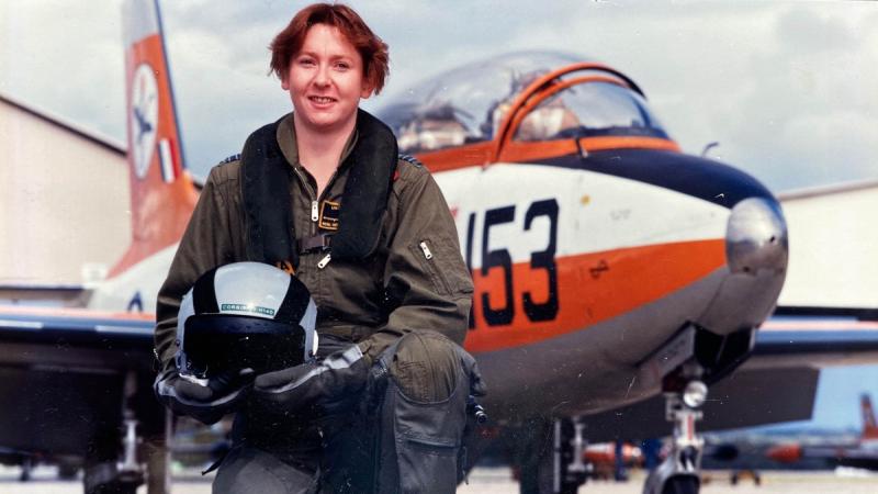 A woman in flight gear holding a helmet kneels in front of a small aircraft.