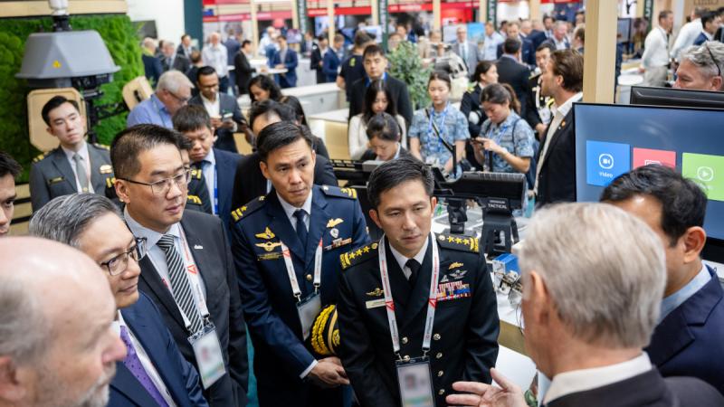 A group of men in suits and military uniform listen to a speaker at a booth in a large convention hall.