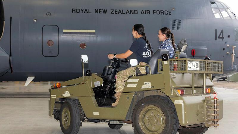 Two aviators seated in a small vehicle approach a large aircraft.