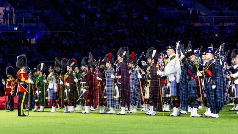 A pipe and drum band marches on the grass field of a stadium.