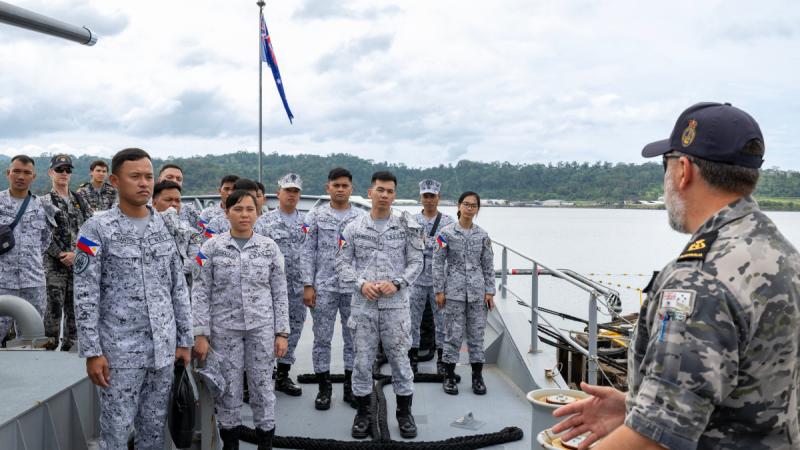 Sailors in fatigues stand on a ship’s deck listening to a man in fatigues speaking.