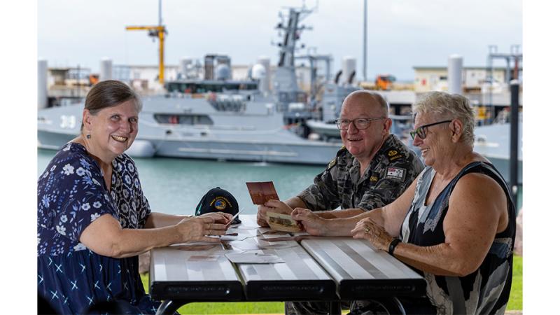 Two civilian women and a military man sit at a table by a shipping dock.