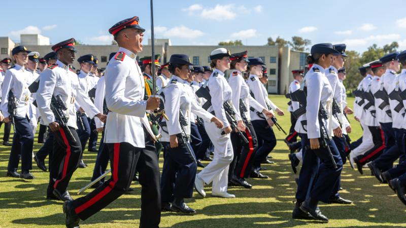 Trainee officers in formal uniform march with machine guns as one walks beside them holding a sword upright.
