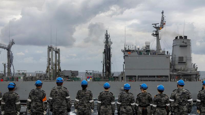 Navy personnel wearing blue protective headwear stand on the deck of a ship.