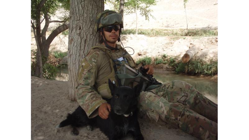 A soldier sits leaning against a tree, with a black dog at his side.