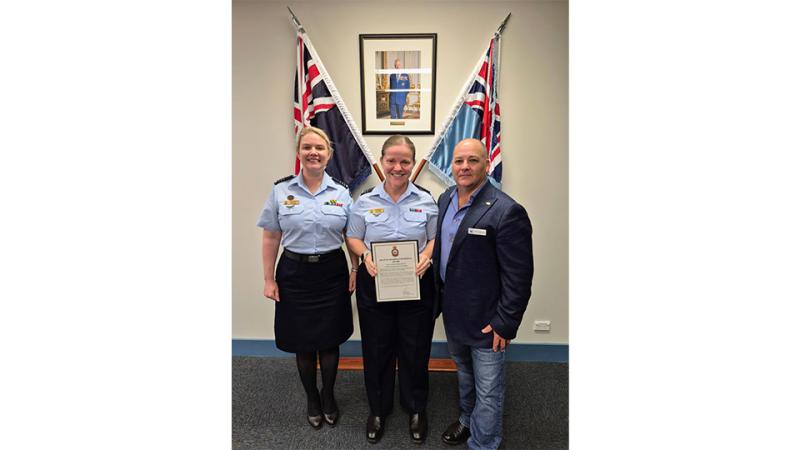 Three people stand indoors as the centre person holds a certificate, with two flags and a framed portrait behind them.