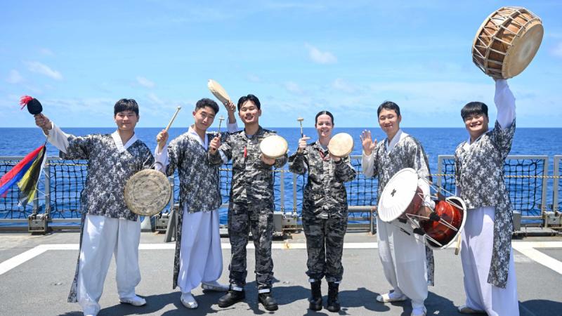 People standing on the deck of a ship holding drums, four in traditional clothing and two in Royal Australian Navy fatigues.