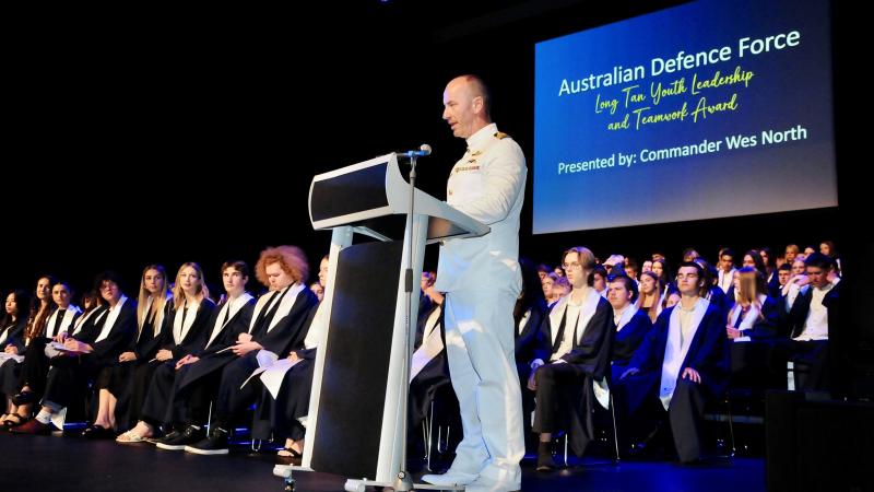 A man in a white uniform speaks at a lectern on a stage with people in graduation robes seated behind him.