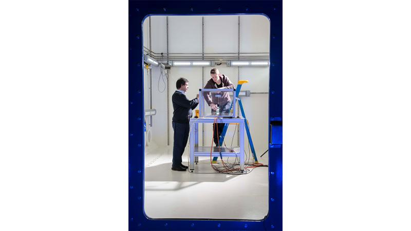 Two men work on equipment inside a white laboratory room viewed through a window of a heavy metal door.