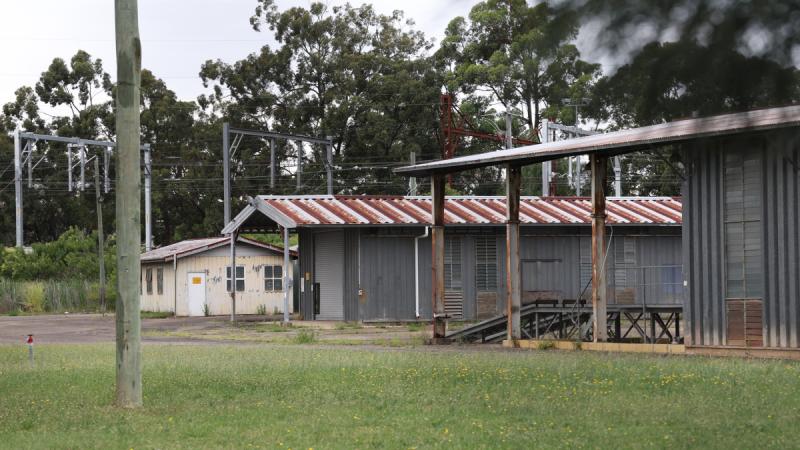 Dilapidated buildings with rusted corrugated iron roofs stand in front of railway overhead lines.