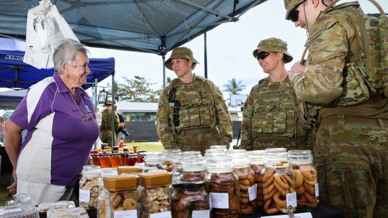 Three women in uniform speak with a stallholder at an outdoor market stall.