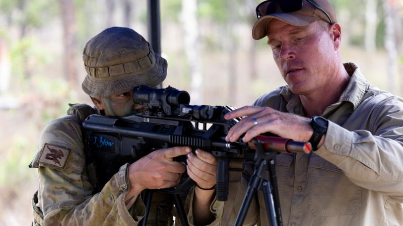 A soldier looks along a weapon mounted on a tripod while a man adjusts the setup.