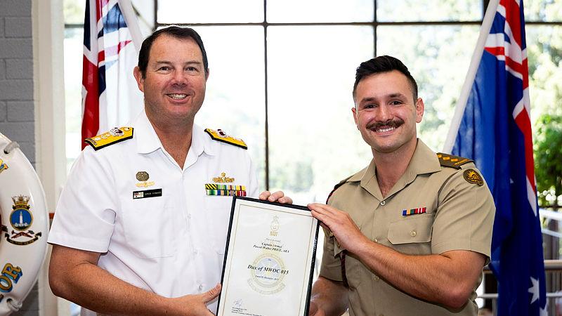A Navy member stands with an Army member, both holding an award certificate.