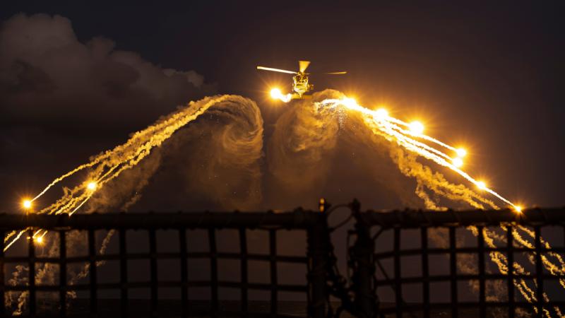 Flares from a helicopter shower into the sea at night, while a rope guardrail blends into the foreground.