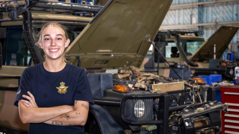 A military woman stands in front of a vehicle.