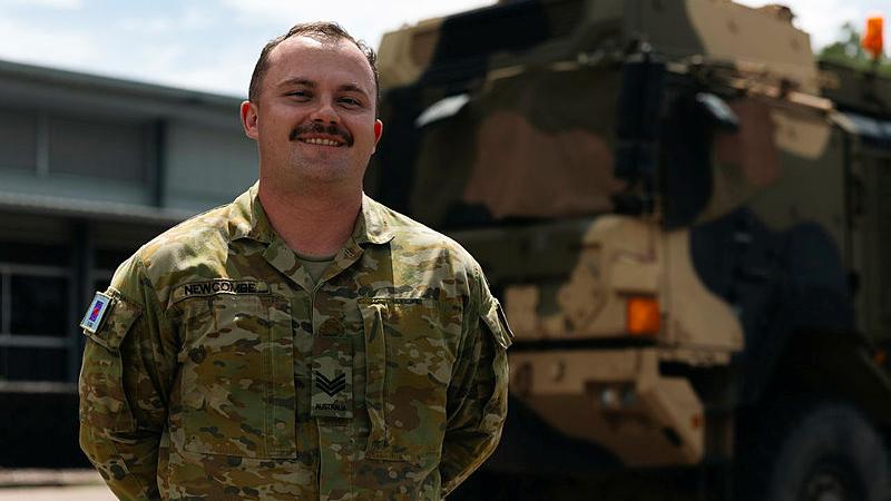 An Army member in uniform stands in front of an Army vehicle.