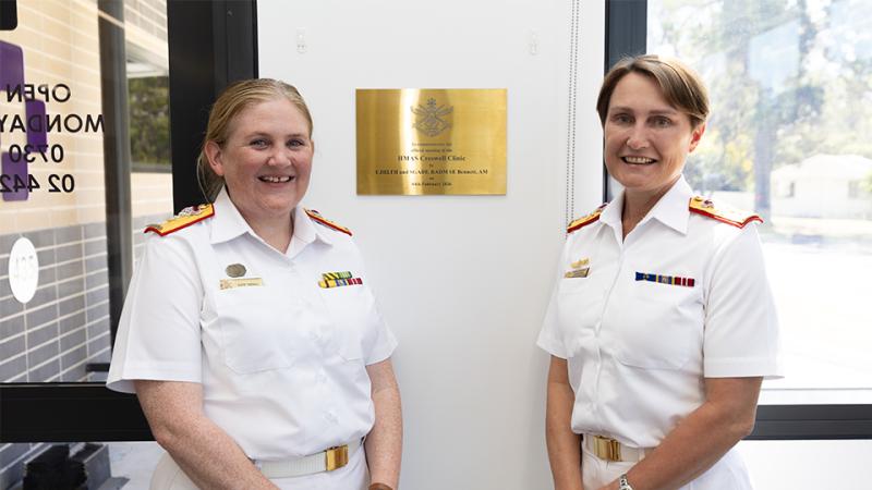 Two women in white uniforms stand indoors beside a mounted plaque on a wall.